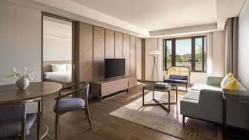 A modern hotel room with a wooden floor, a flat-screen TV on a wooden console, a sofa, a dining table with chairs, and a large window offering a view of palm trees.