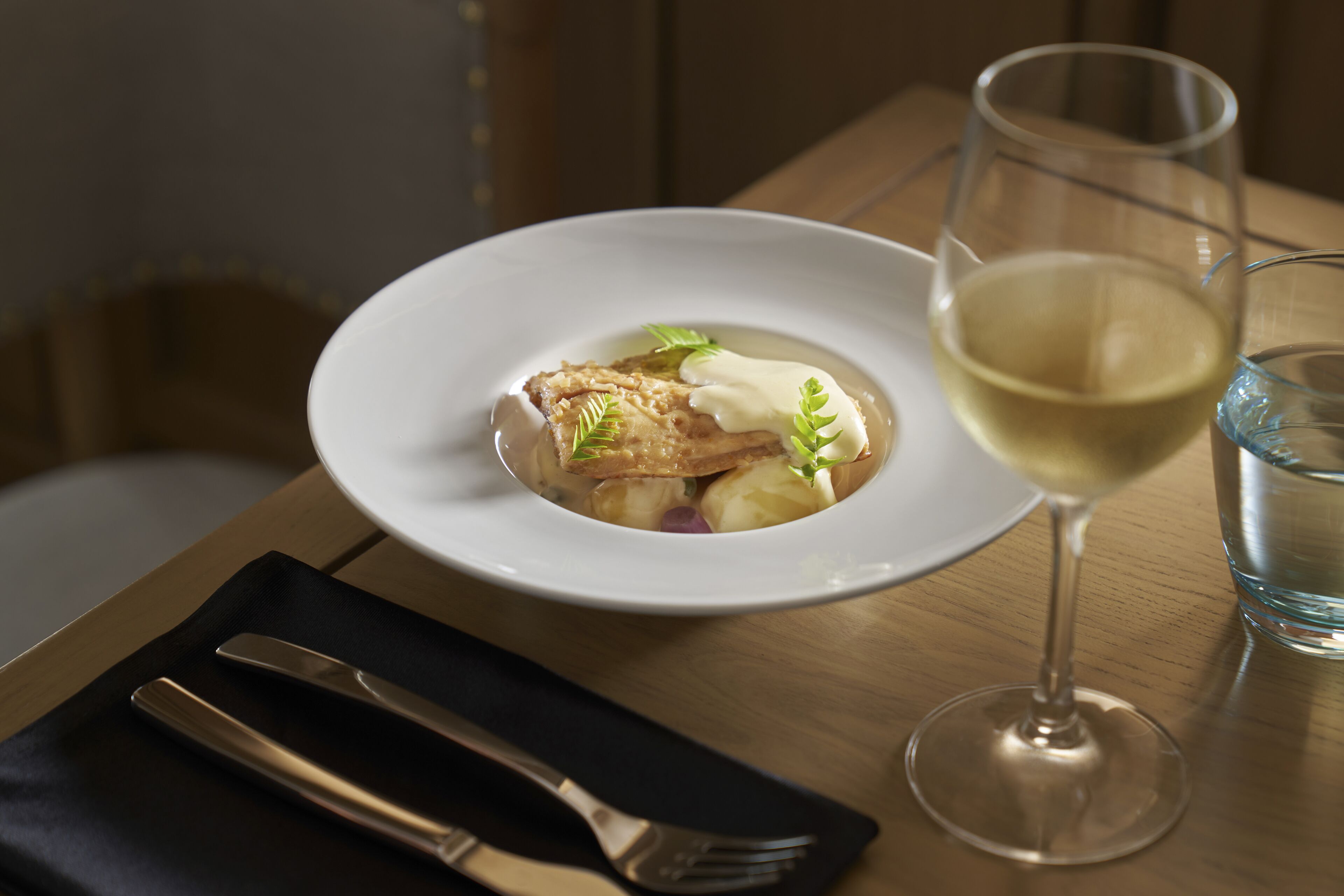 Een wit bord met een gerecht en een glas witte wijn op een houten tafel.