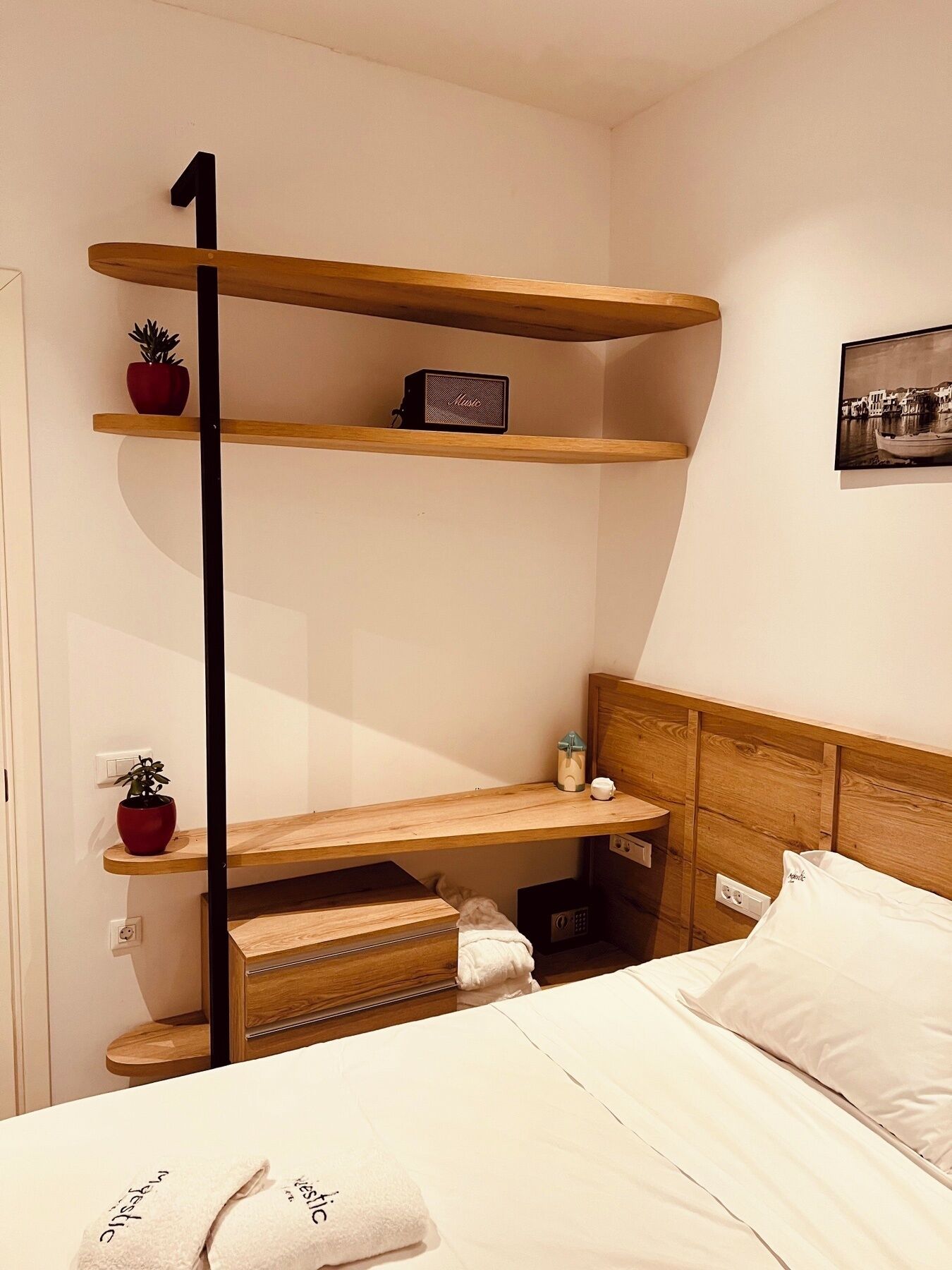 Un dormitorio con una cama, cabecera de madera, mesita de noche, estante con una planta, un altavoz pequeño y un cuadro en la pared.