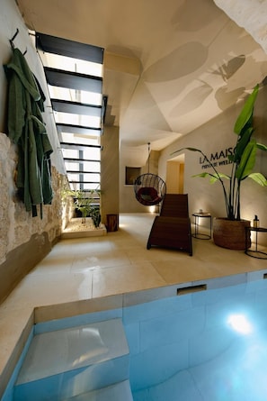 Un espace piscine intĂ©rieur moderne, dotĂ© dâun fauteuil suspendu, dâun escalier et dâune enseigne indiquant : « LA MAISON, ESPACE DĂDIĂ AU BIEN-ĂTRE PRIVĂ ».