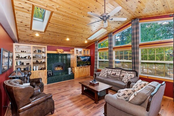 A cozy living room with a fireplace, wooden ceiling, and large windows.