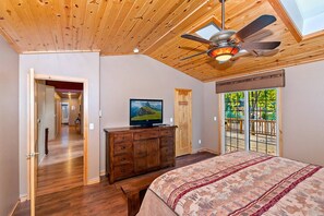 A bedroom with a wooden ceiling, a bed with a patterned bedspread, a dresser, a television, and a view of the outdoors through a sliding door.