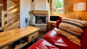 Interior acogedor de una cabaña con chimenea de piedra, escalera de madera, un sofá de cuero rojo y una mesa de madera con un juego de Scrabble.