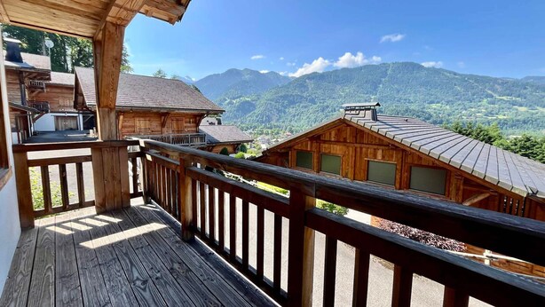 Un balcón de madera con vistas a montañas y un edificio con tejado inclinado.