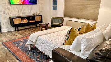 A bedroom with a bed, a sofa, a TV, a small table, and a window with blinds.