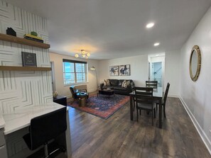 Un salon moderne avec un canapé, une table à manger et une table basse. Une fenêtre offre une vue sur les bâtiments à l’extérieur.