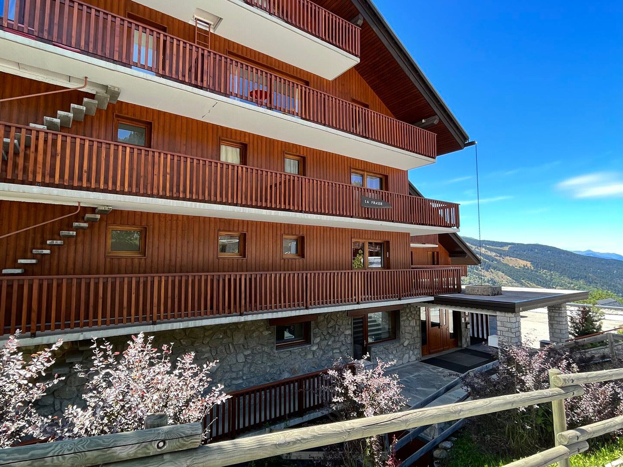 A multi-story wooden building with balconies, stone foundation, and a view of mountains.
