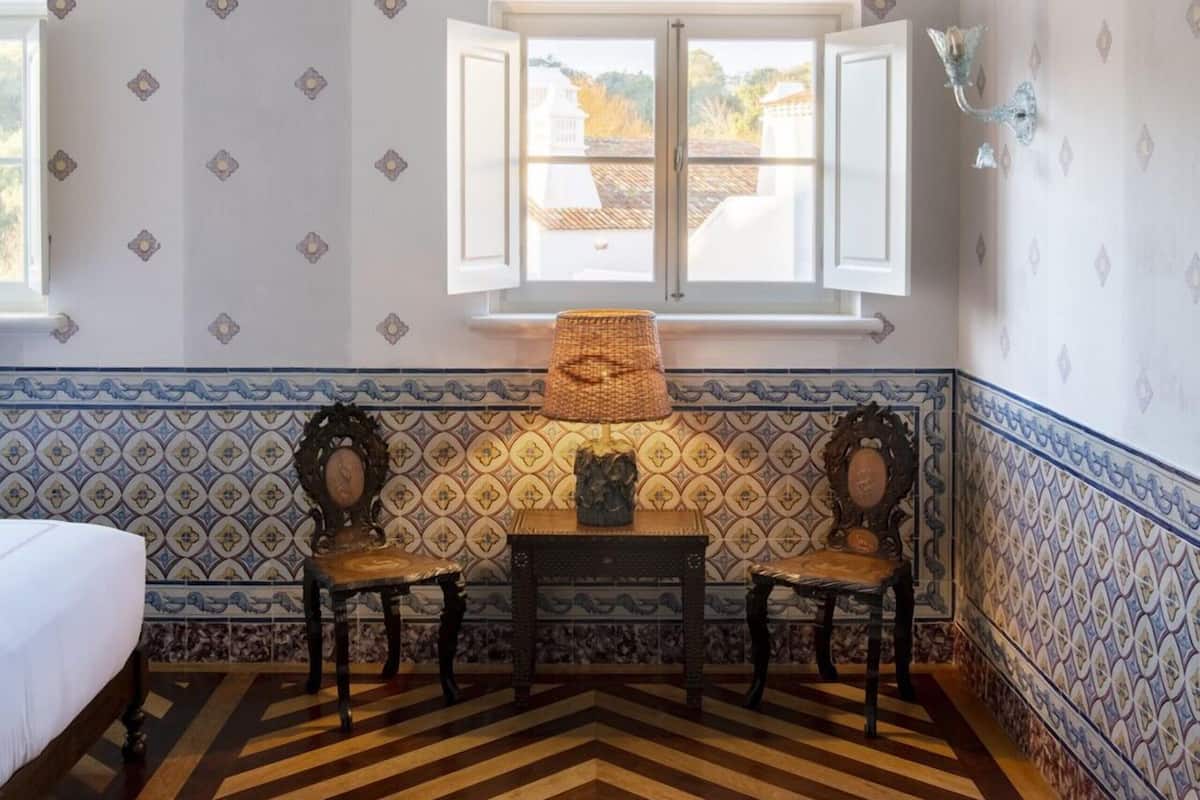 A room with a patterned floor, a bed, two chairs, a table, and a window with open shutters.