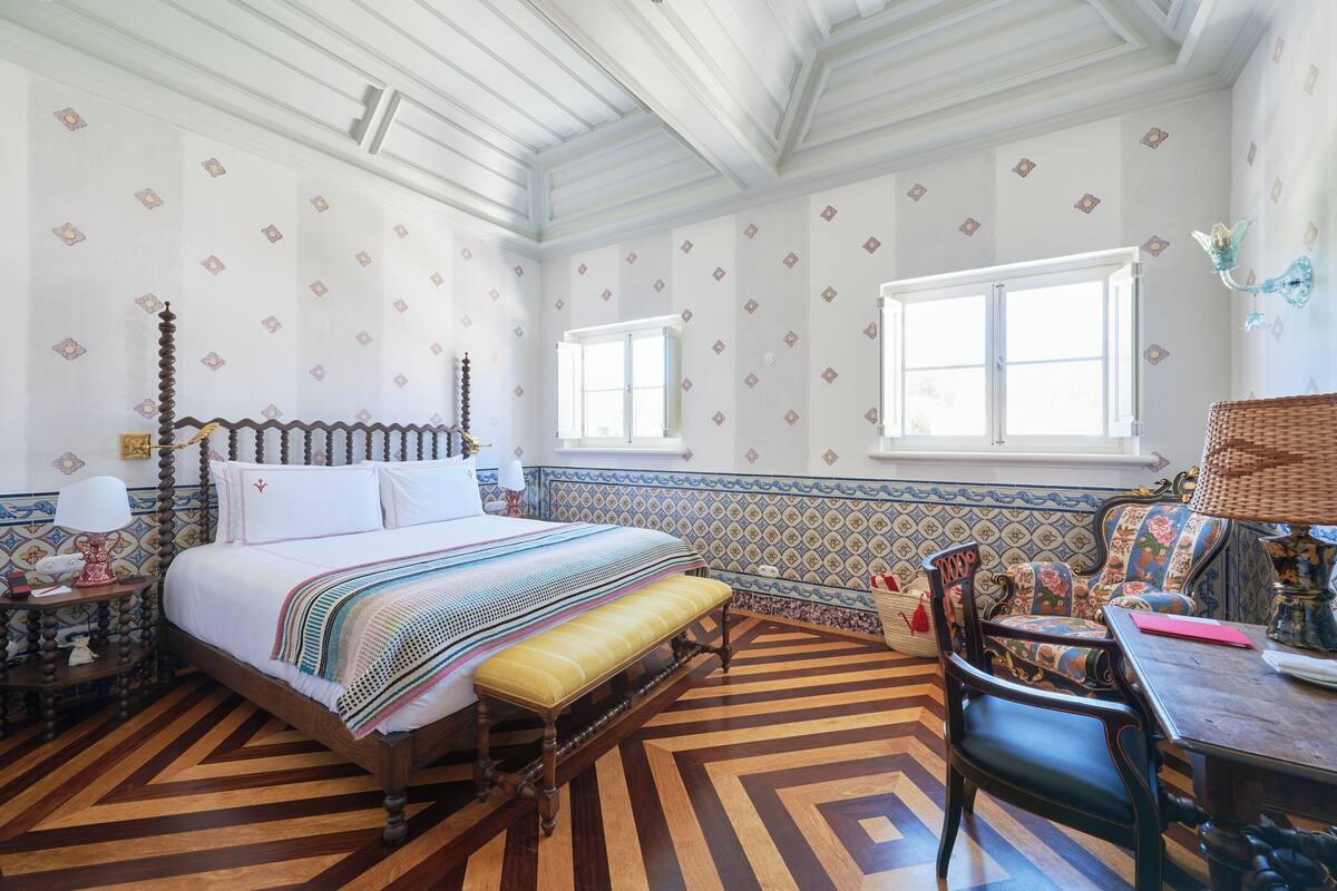 A traditional room with a large bed, a wooden bench, a chair, a table, and a window.