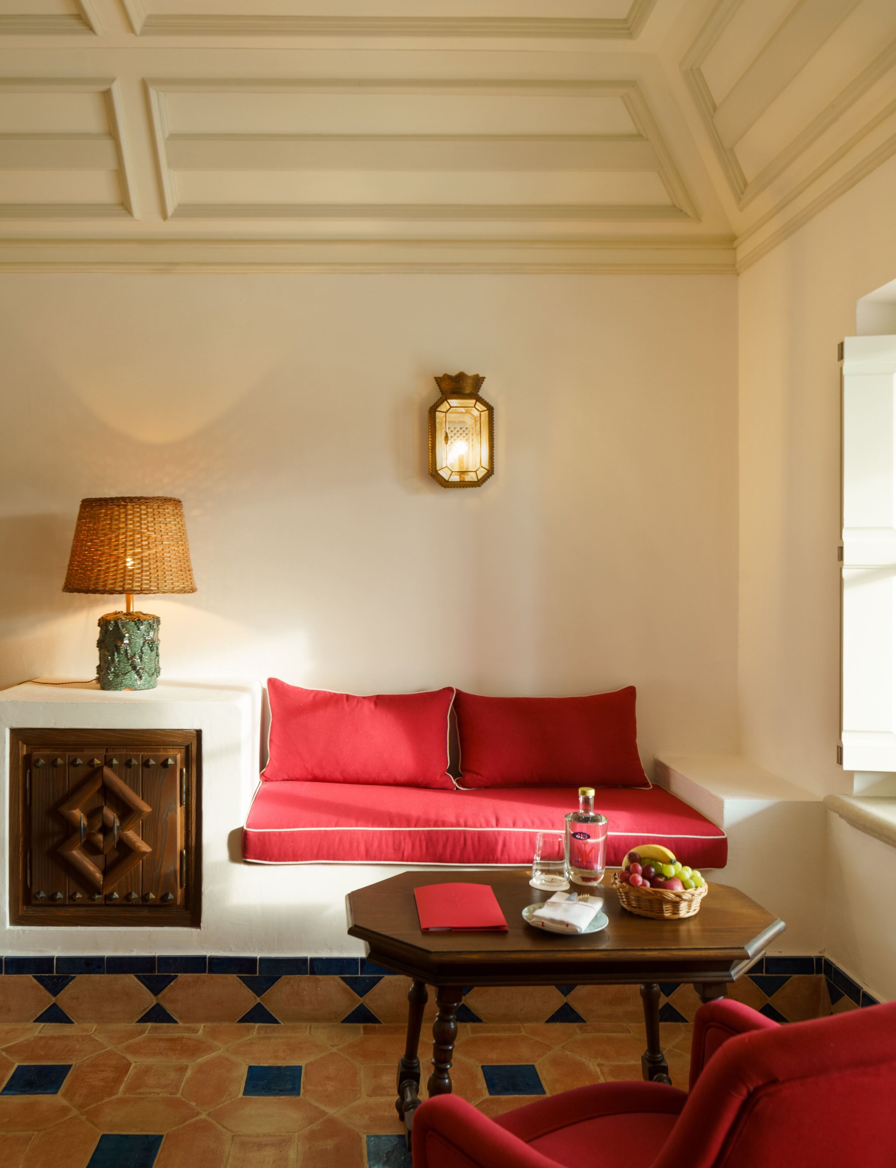 Una habitación con un sofá rojo, una mesa de madera con un vaso, una botella y una cesta de fruta.