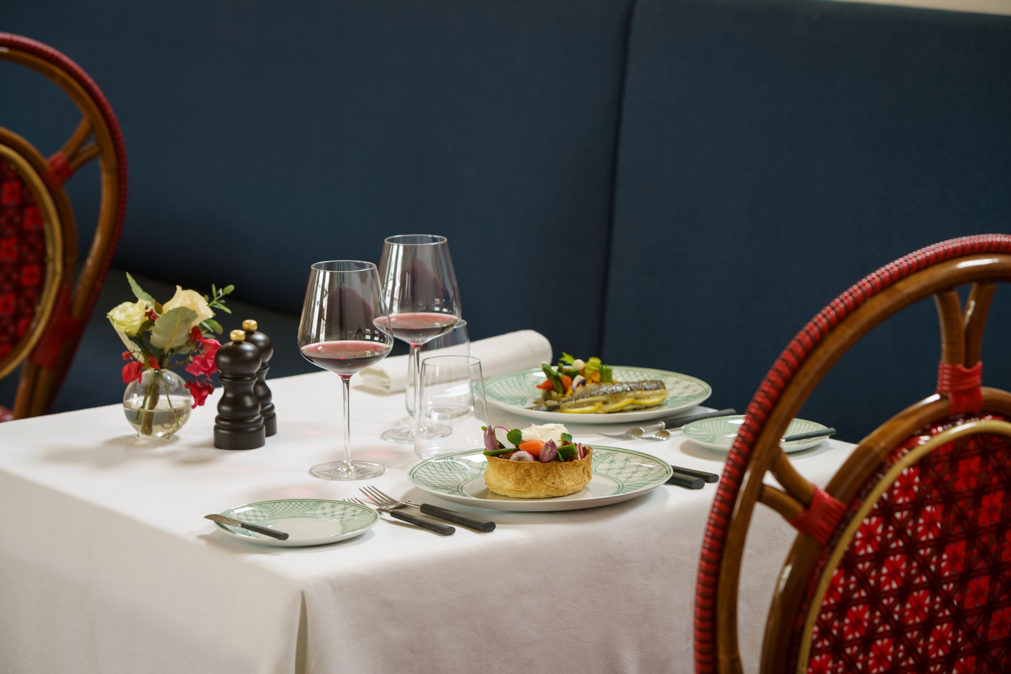 Una mesa de comedor dispuesta con platos, cubiertos, un florero con flores y dos copas de vino tinto.