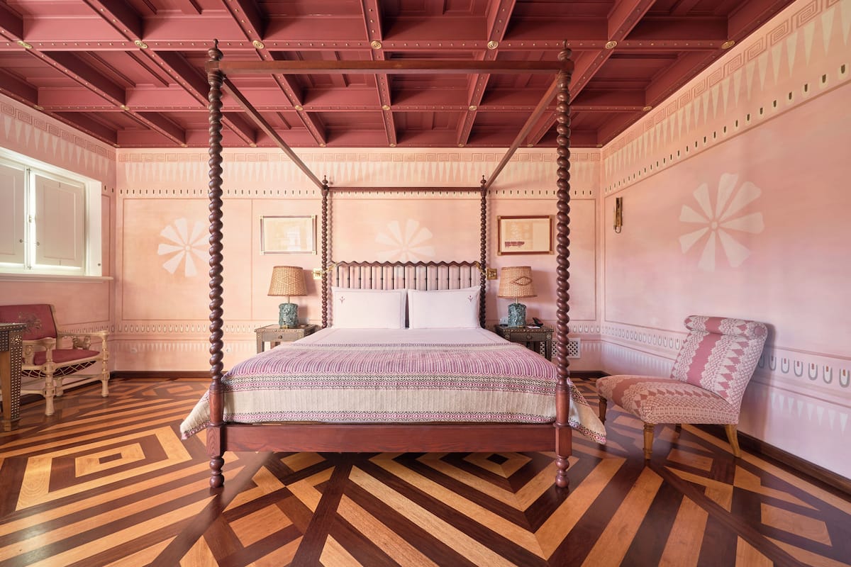 A four-poster bed with a patterned bedspread, a wooden floor with a geometric design, and a pink and white color scheme.