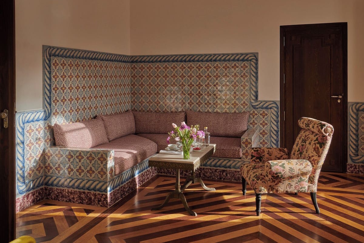 A room with a patterned sofa, a floral armchair, a small table with a vase, and a wooden floor with a chevron pattern.