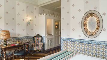 A room with patterned wallpaper, a decorative mirror, a wooden table with a lamp, and a chair.