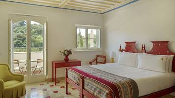 A bedroom with a bed, a chair, a table, a window, and a view of greenery outside.