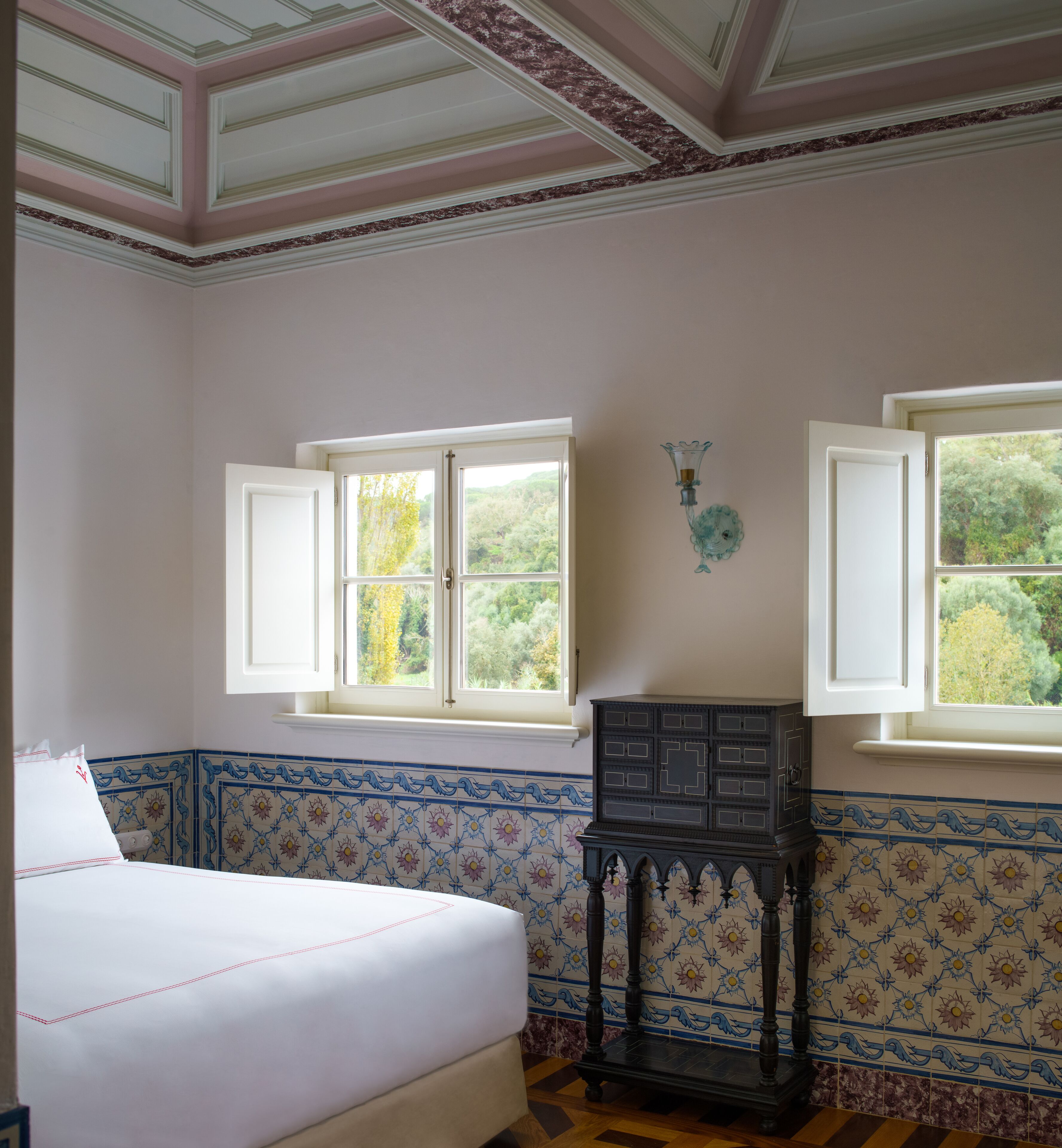 Quarto com cama, duas janelas, luminária na parede e um armário decorativo.