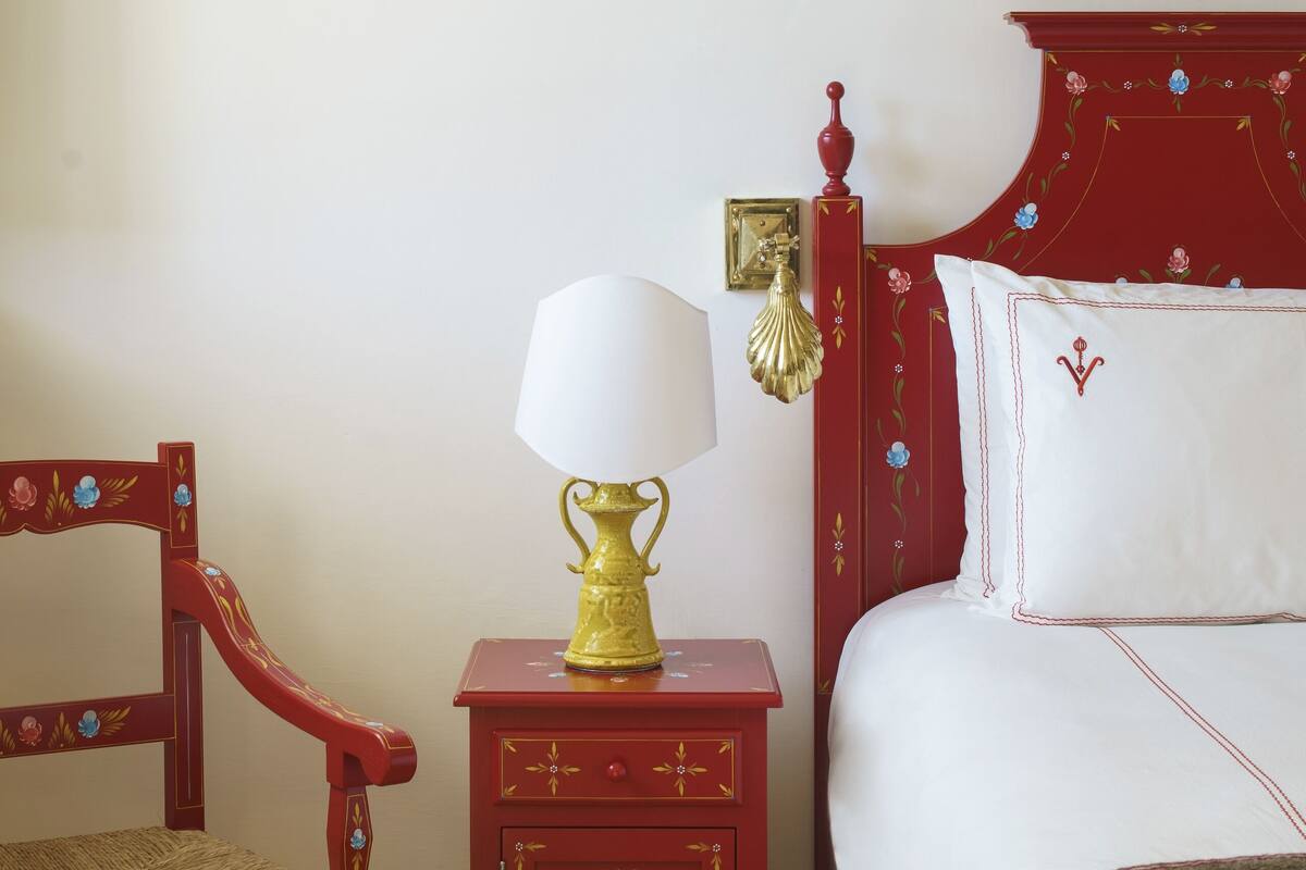 A red wooden bed with white bedding, a matching nightstand with a lamp, and a wooden chair with a woven seat.