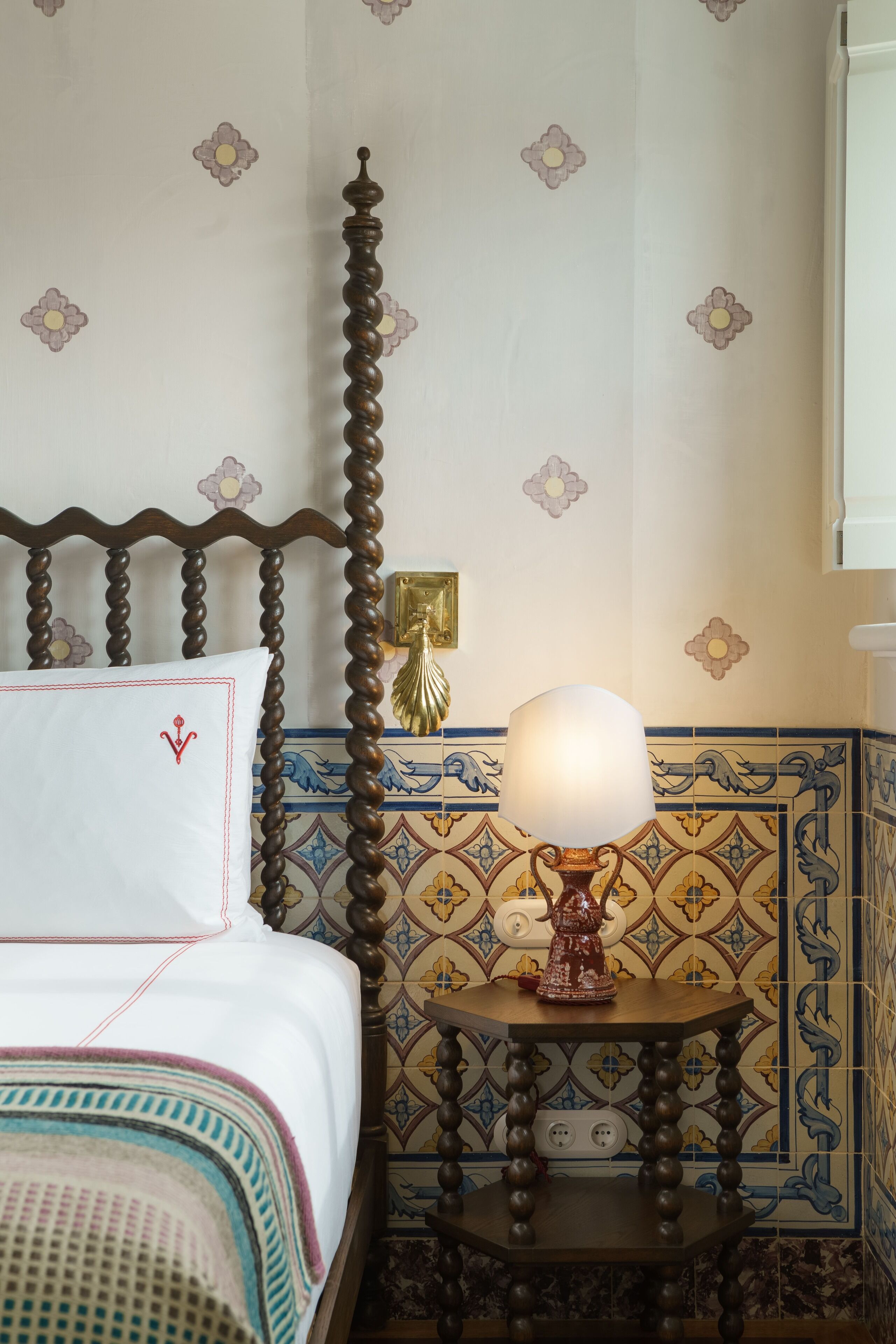 A bed with a wooden headboard, a bedside table with a lamp, and a wall with floral and geometric tile patterns.