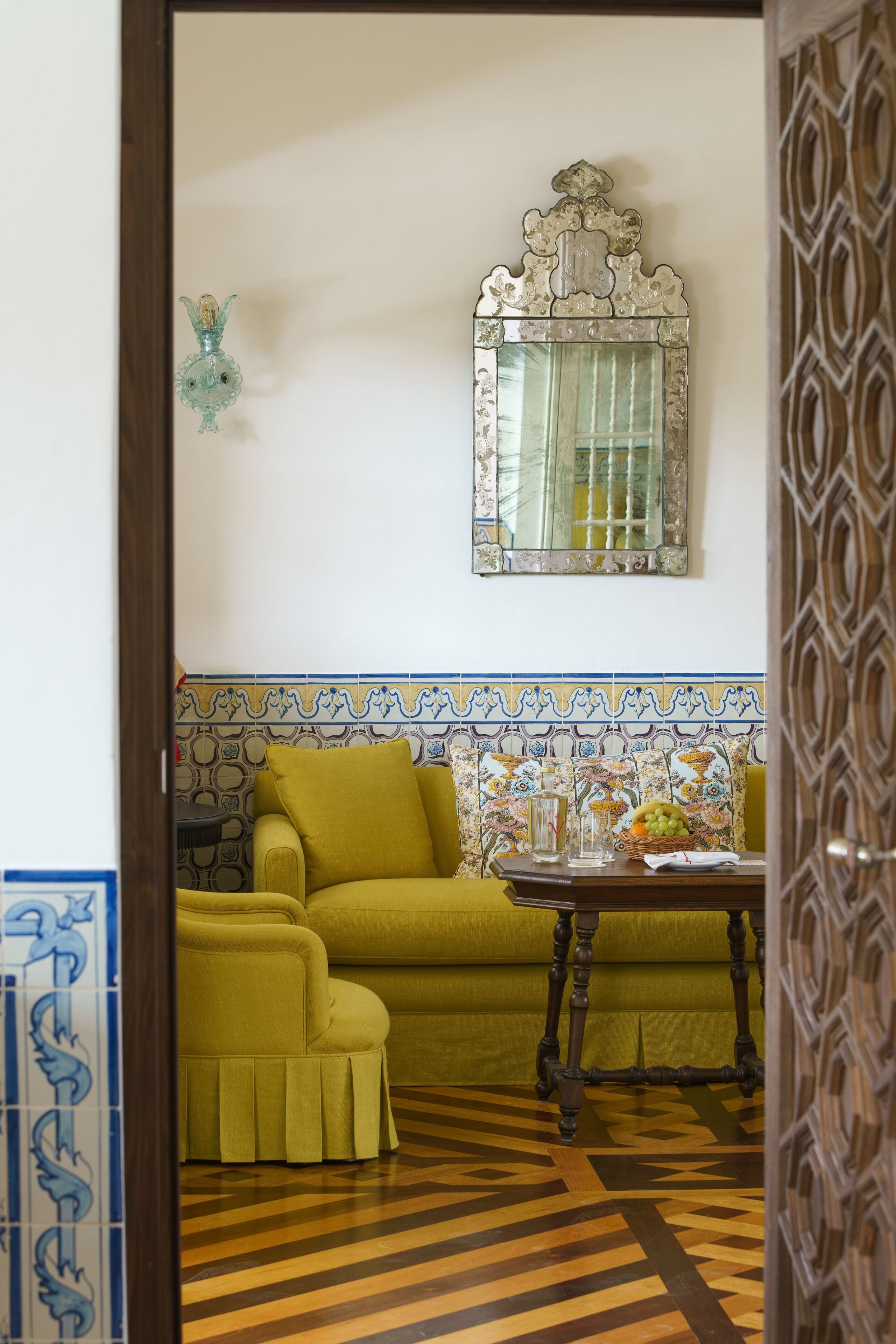 A room with a patterned tile backsplash, a green sofa, a wooden table with a vase, and a mirror with an ornate frame.