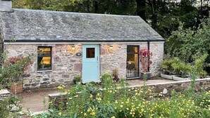 A stone cottage with a light blue door, surrounded by a garden with yellow flowers. - Charming stone Bothy at Loch Lomond (Muirlands, Arden, Alexandria, Luss,)