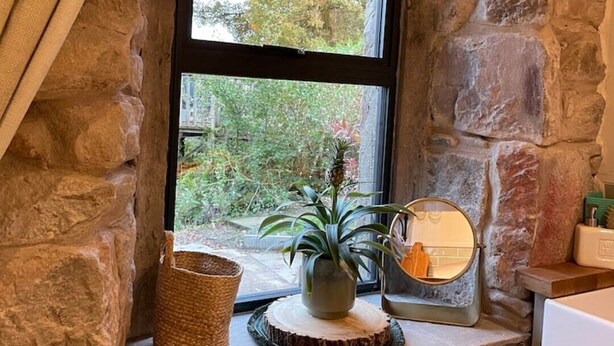 A stone-walled bathroom with a window, a potted plant, a woven basket, and a round mirror. - Charming stone Bothy at Loch Lomond (Muirlands, Arden, Alexandria, Luss,)