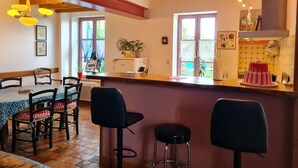 A cozy kitchen with wooden beams, a dining area with patterned tablecloths, and a view of the garden through the windows. - Charming Cottage: Enclosed Garden, Close to Loire Valley, Sleeps 6 (Beaulieu-sur-Loire)