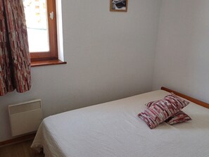 A single bed with a patterned pillow, a window with curtains, and a wall-mounted air vent.
