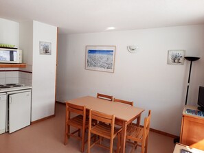 A compact kitchen with a stove, microwave, and refrigerator. There is a wooden dining table with four chairs. The walls are plain with two framed pictures.