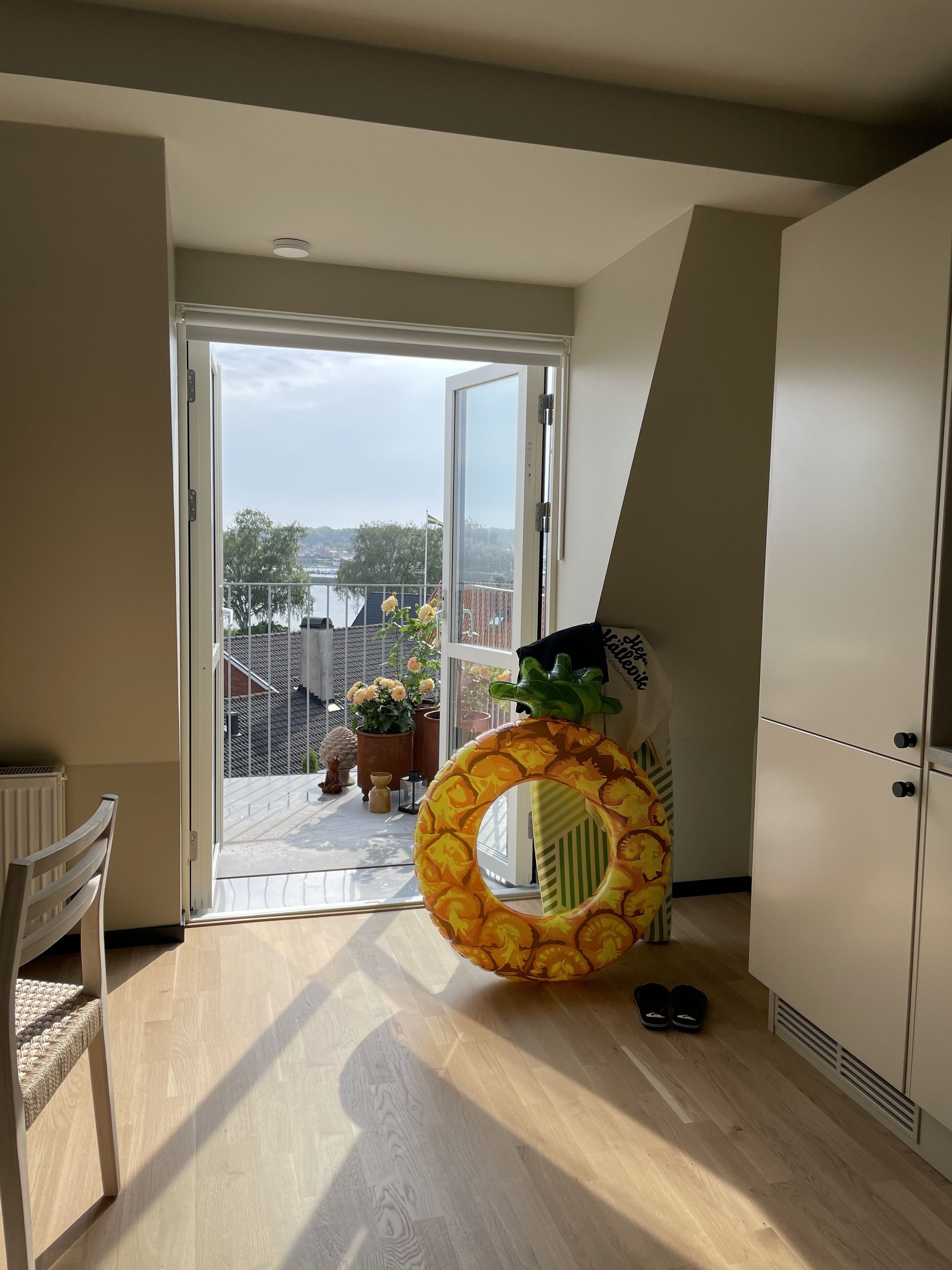 Une pièce dotée d’une porte-fenêtre coulissante qui donne sur un balcon offrant une vue sur un plan d’eau et des arbres. On y trouve un plancher en bois, une chaise blanche et un grand anneau décoratif posé sur le sol.