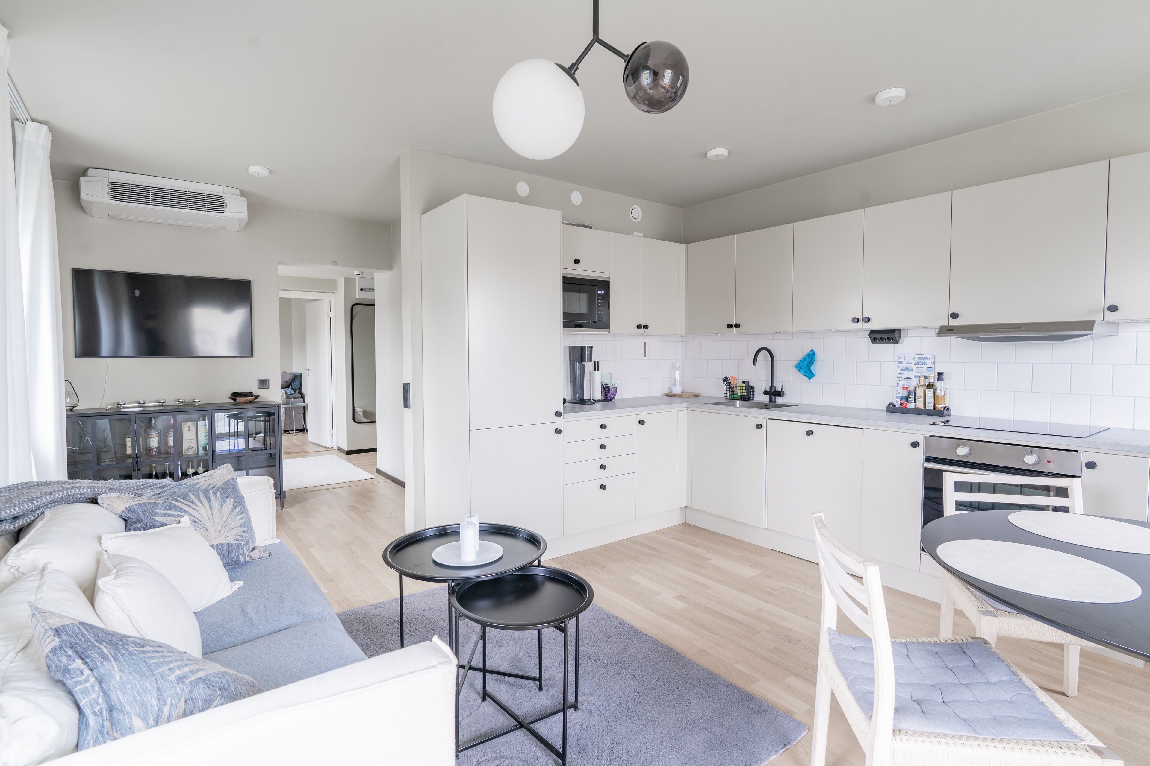 Cozinha moderna com armários brancos, área de jantar com mesa e cadeiras, e sala de estar com sofá e mesa de centro.