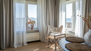 Una habitación con vista al mar, una silla, una mesa con individuales tejidos y un jarrón con flores secas.