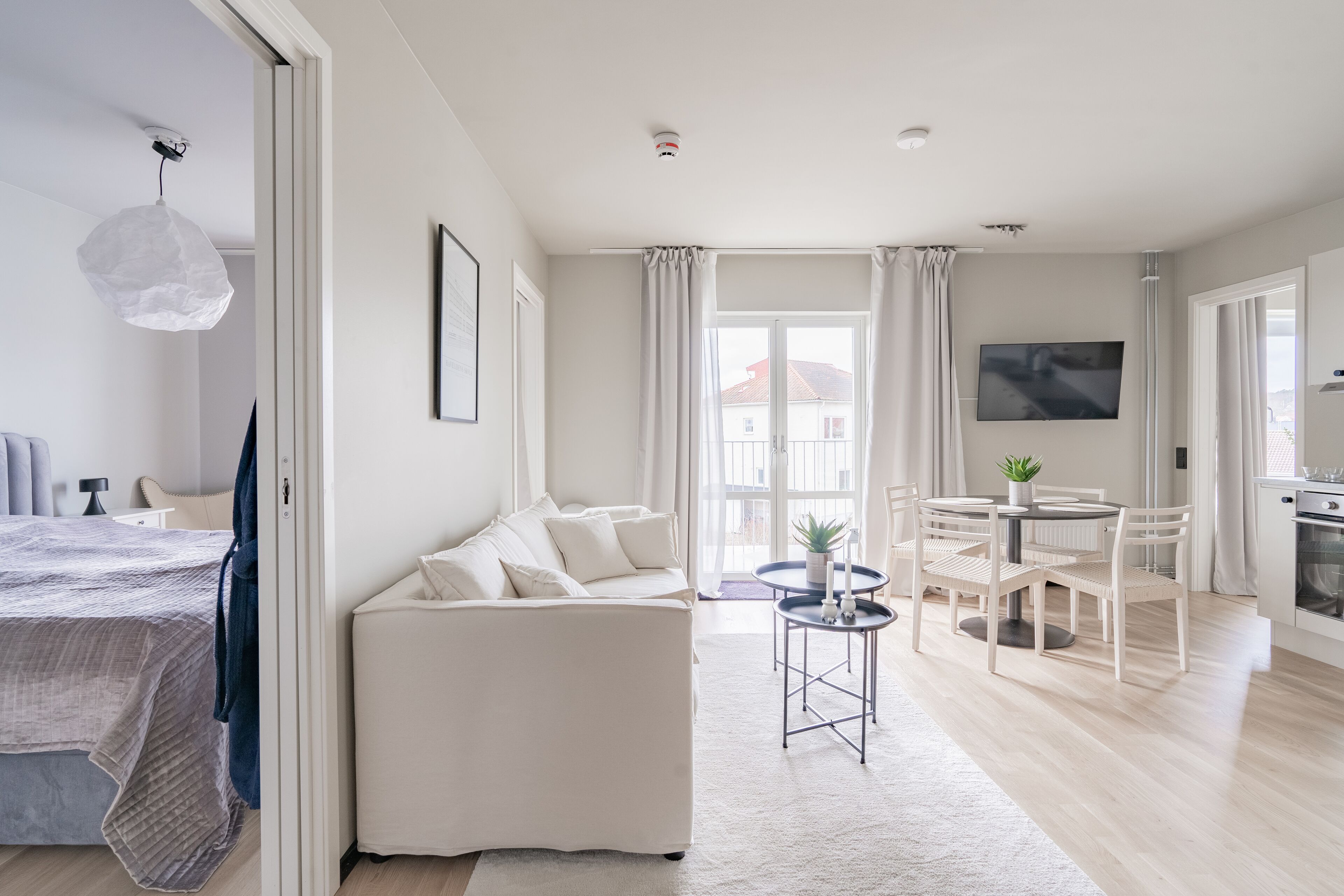 A modern, well-lit living room with a sofa, dining table, and a TV mounted on the wall.