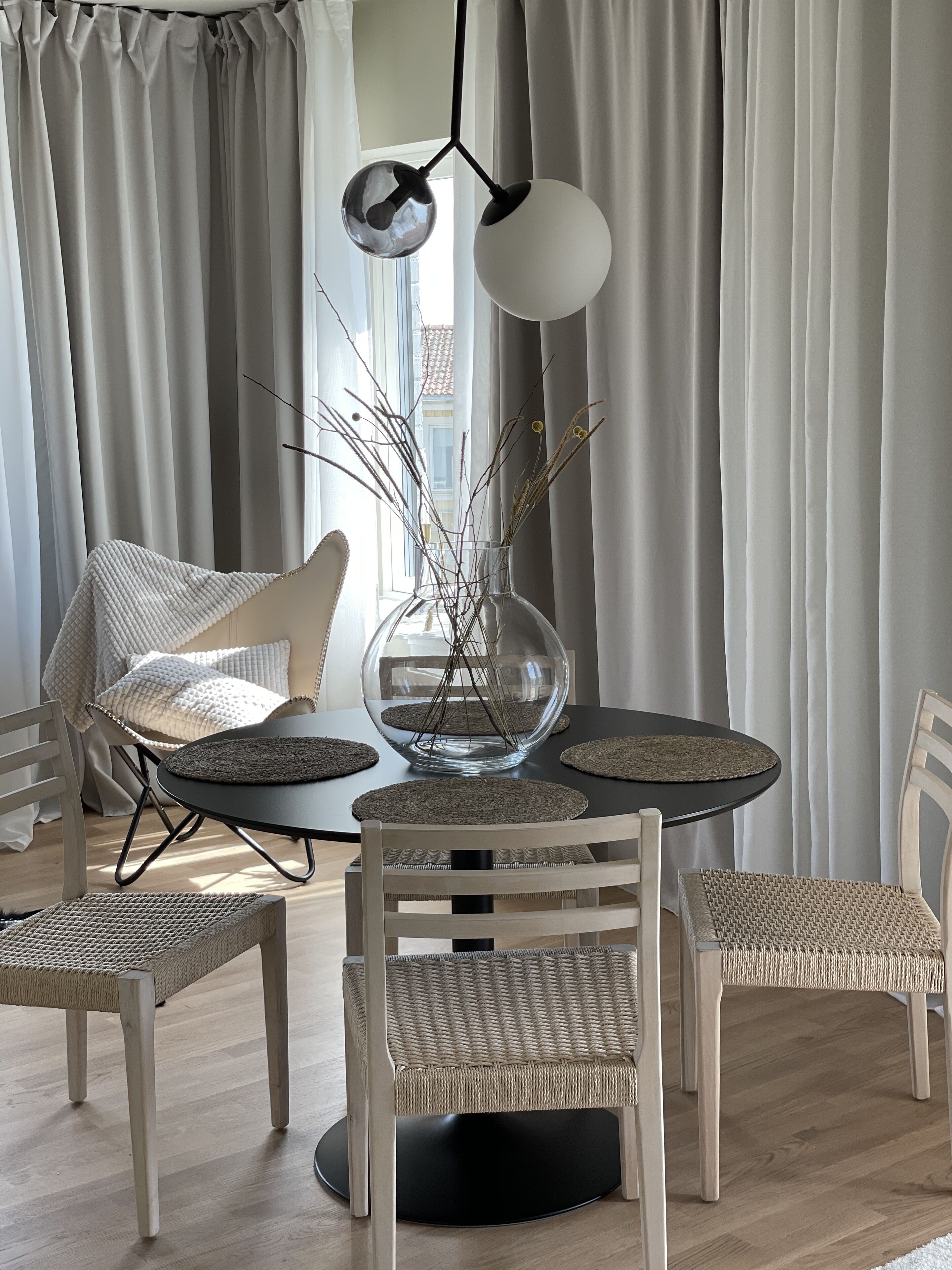 A dining area with a round table, four wicker chairs, a glass vase with branches, and a hanging lamp.