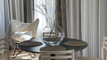 A dining area with a round table, four wicker chairs, a glass vase with branches, and a hanging lamp.
