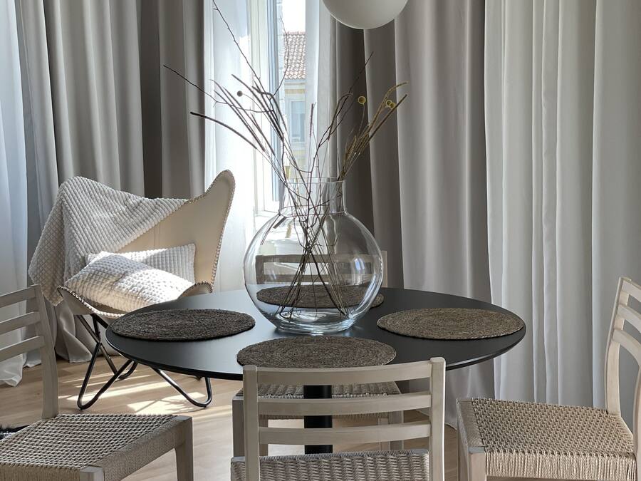 A dining area with a round table, four wicker chairs, a glass vase with branches, and a hanging lamp.