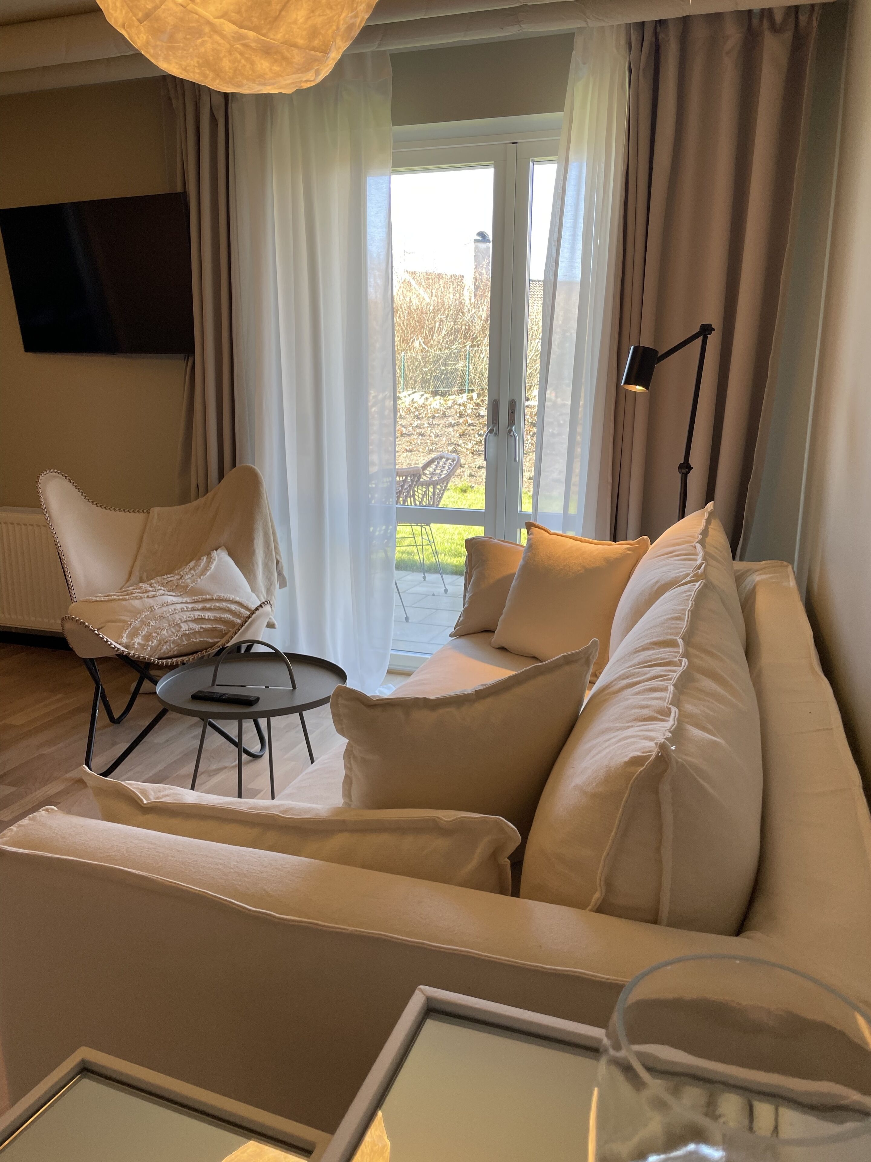 Un salon moderne avec un canapé blanc, un fauteuil et une petite table ronde. Une porte vitrée donne accès à un balcon d ’où l’on peut admirer un jardin.
