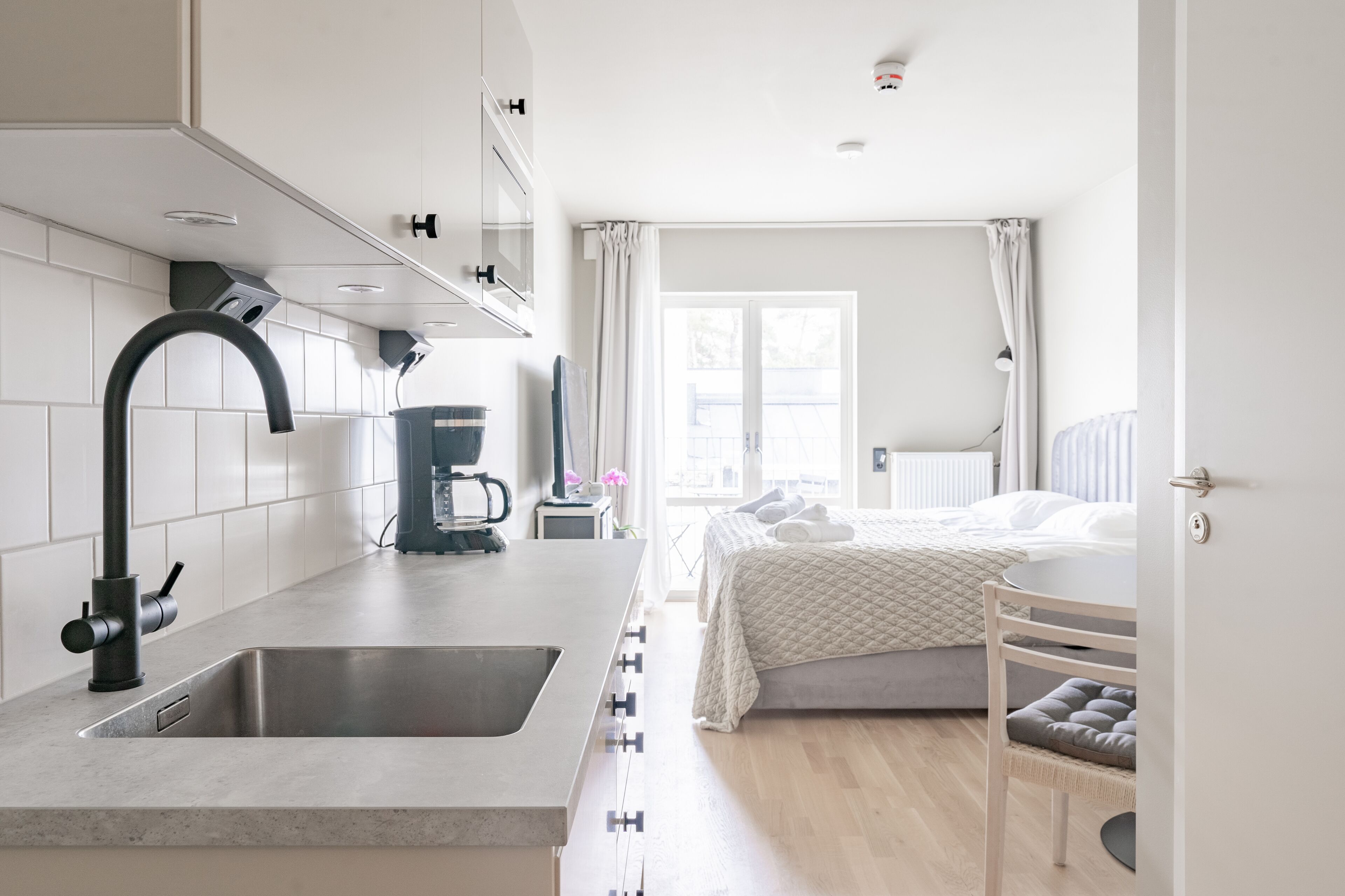 A modern kitchen with a built-in sink and a bed with a bedside table in the adjacent room.