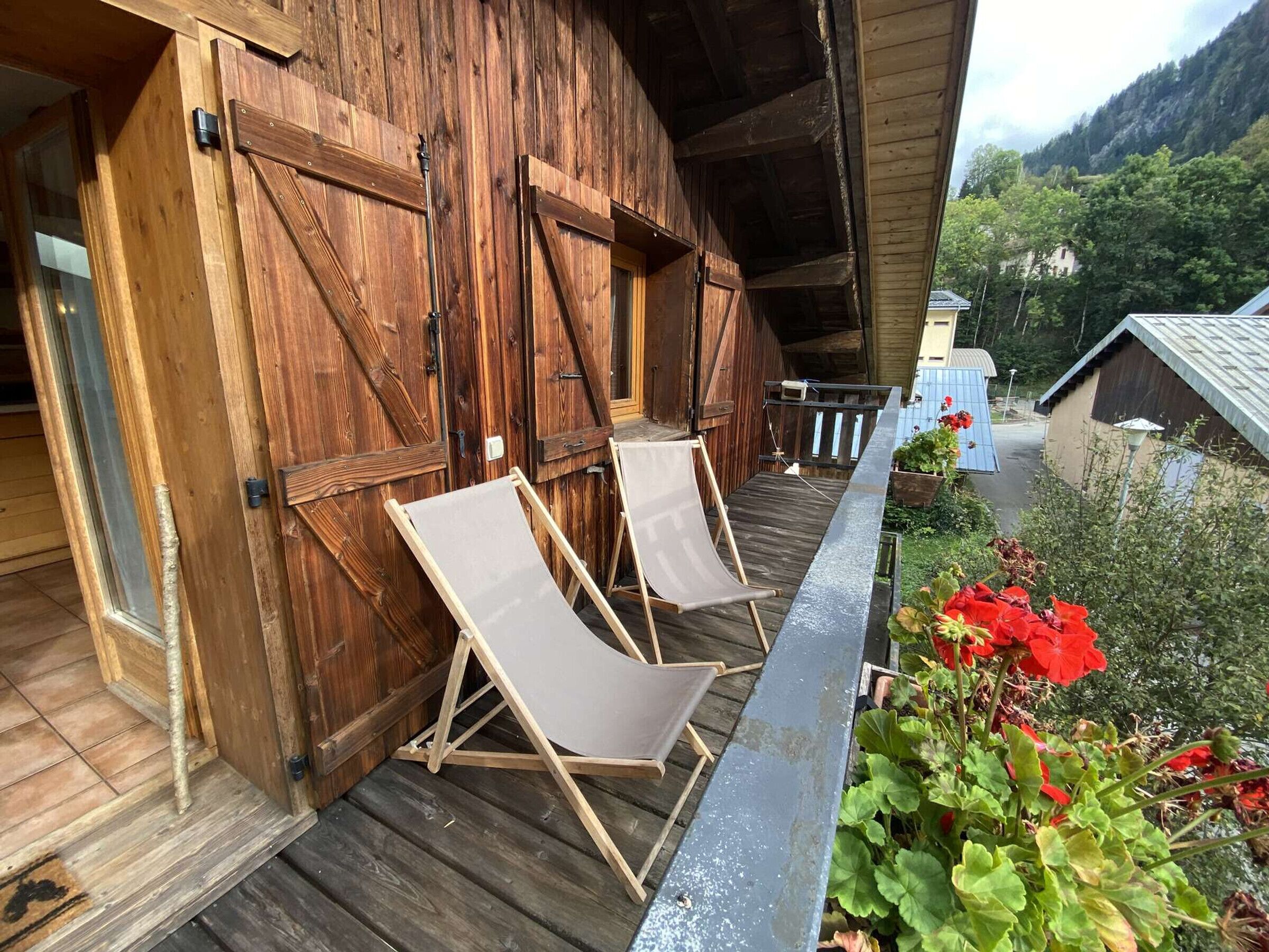 Eine Holzhütte mit einem Balkon, zwei Klappstühlen und einem Garten mit roten Blumen.