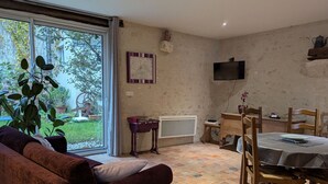 Ein gemütliches Wohnzimmer mit einer Couch, einem Esstisch mit Stühlen, einem an der Wand montierten Fernseher und einem großen Fenster mit Blick auf den Garten.