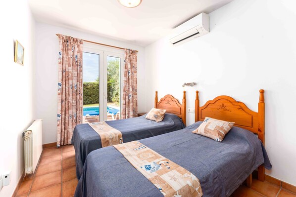 A bedroom with two beds, a window with curtains, an air conditioning unit, and a view of a swimming pool. - Cosy detached villa with private pool near the beach (Binibequer)