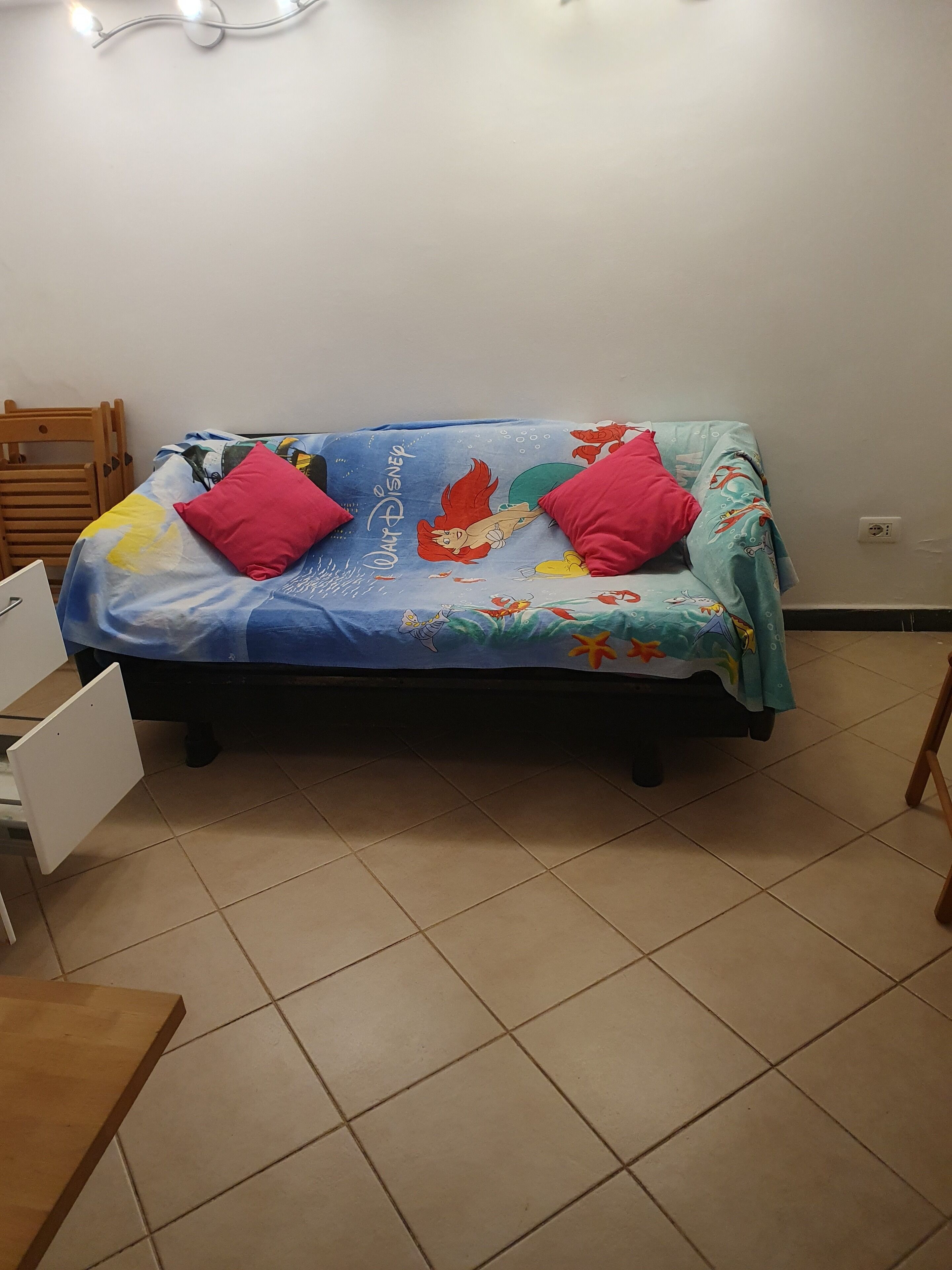 Un lit avec une couvre-lit aux motifs de Disney, deux oreillers rouges et une chaise en bois.