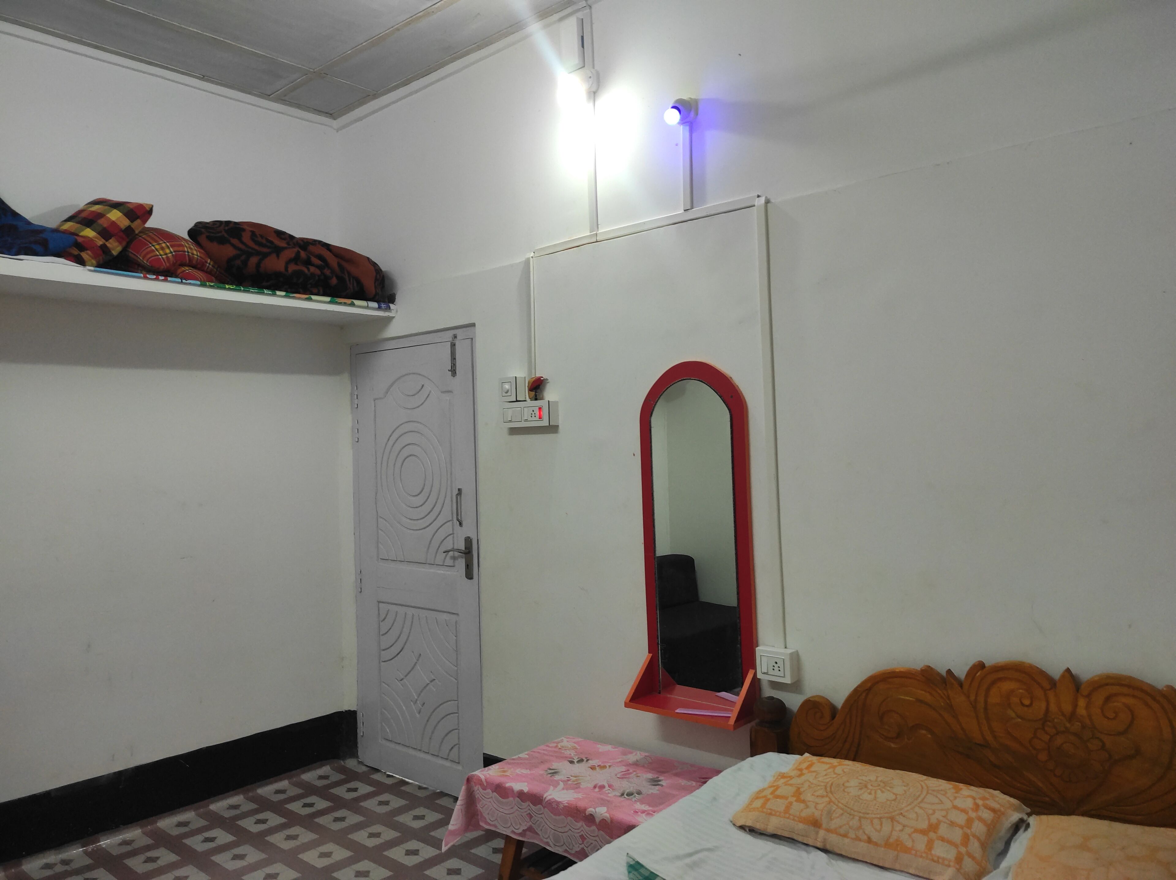 Quarto com cama, uma mesinha, espelho, prateleira com almofadas e porta.