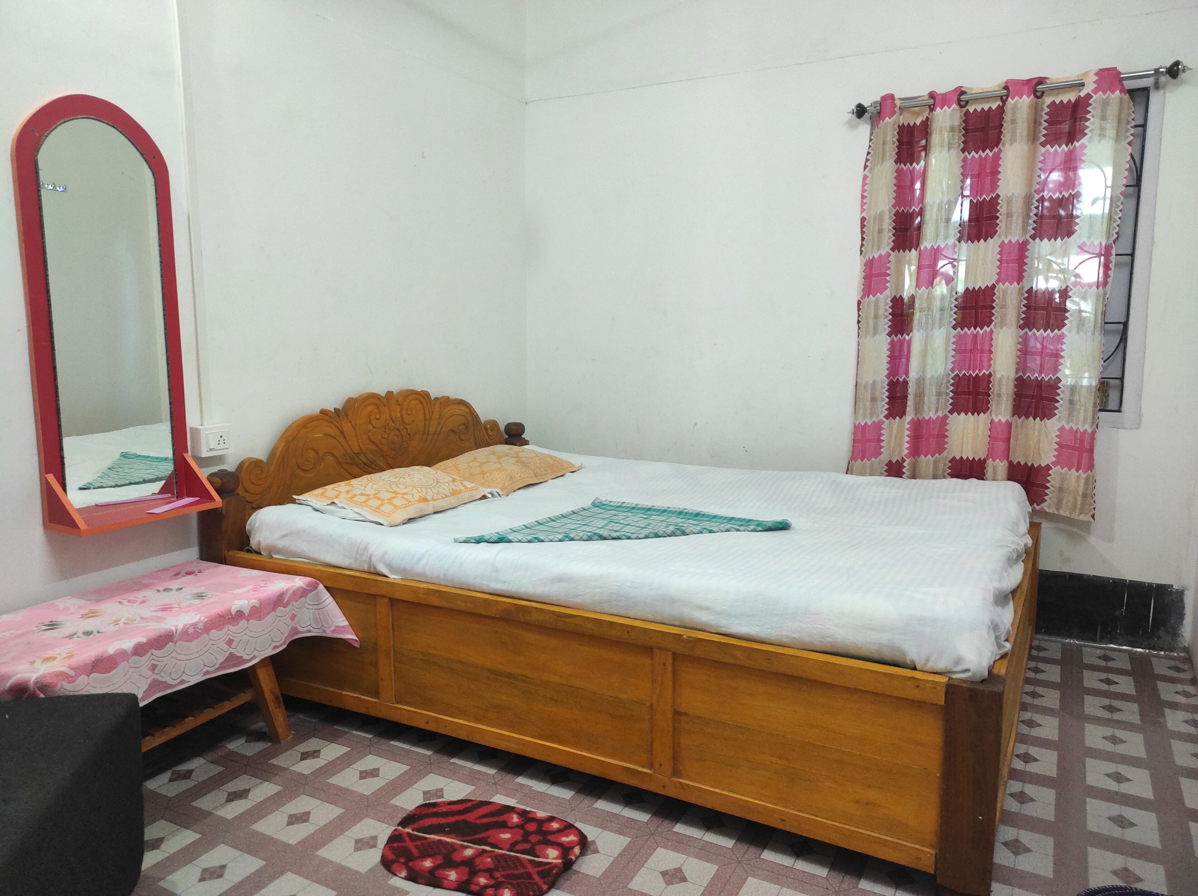 A single-bed room with a wooden headboard, a bedside table, a mirror, and a window with patterned curtains.