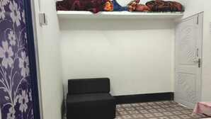 A small room with a bed, a black sofa, a patterned floor, a wardrobe, and a shelf with blankets.