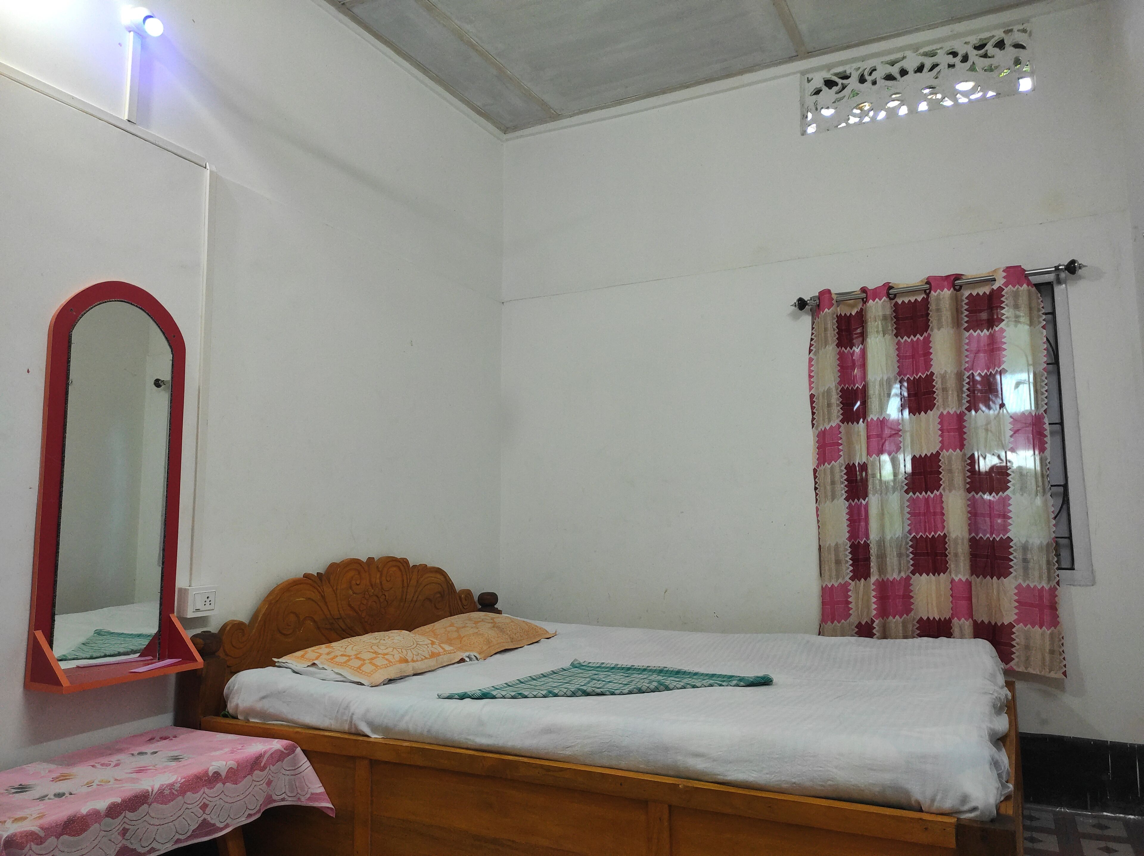 A bedroom with a wooden bed, a mirror, a bedside table, and a window with patterned curtains.