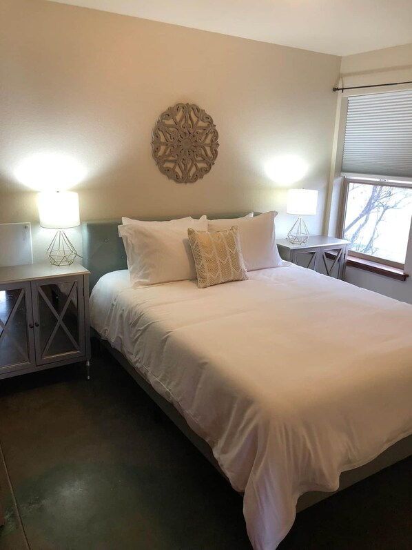 Una cama bien hecha con sábanas blancas y almohadones decorativos, dos mesitas de noche con lámparas y un adorno en la pared sobre la cama.