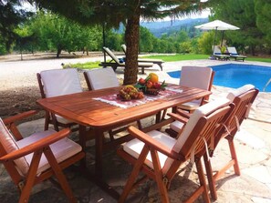 A wooden table with white cushioned chairs, a fruit basket, and a pool in the background.