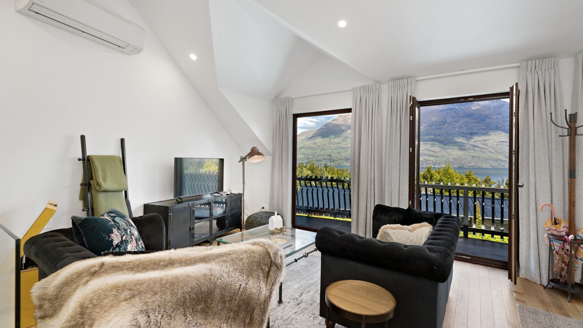 A modern living room with a sofa, a coffee table, and a TV. There's a balcony with a view of mountains and a lake.