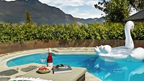 A poolside setting with a swan float, a bottle of wine, a glass, and a book on a table. - Alpine Cottage | Swimming pool | Lake and Mountain Views (Queenstown)