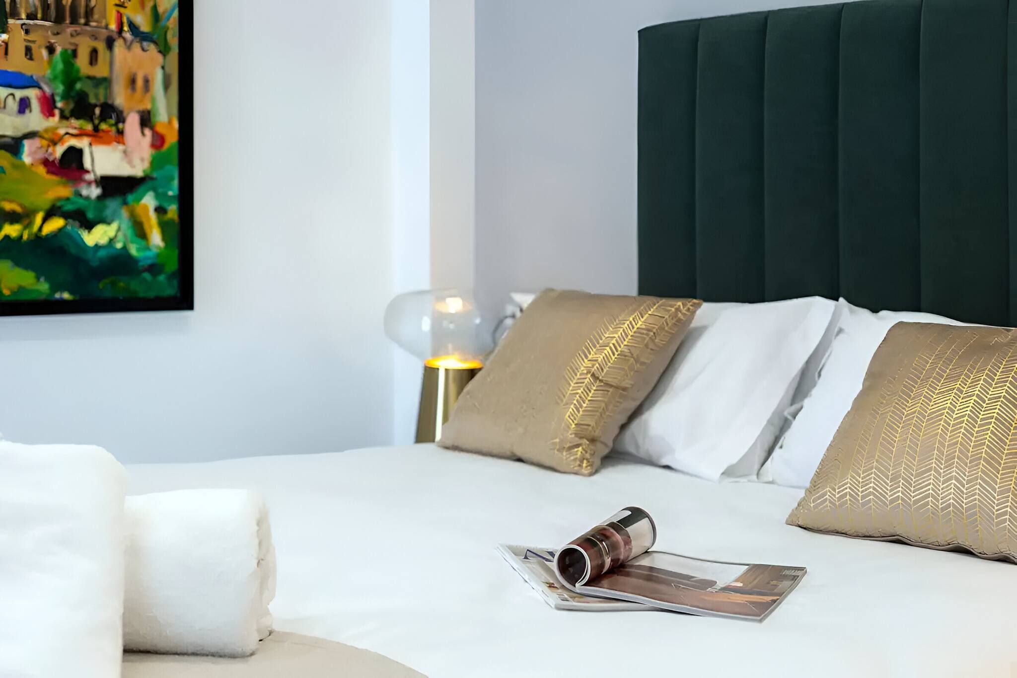 Una cama bien hecha con almohadas blancas y doradas, una revista y una lámpara de vidrio sobre una superficie blanca.